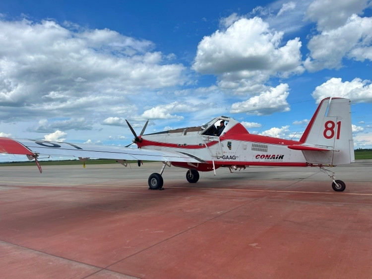 A white and red airtanker with the number 81 painted on its tail sits on an airport tarmac under a partly cloudy sky. The plane is labeled "Conair". The pilot is visible in the cockpit.