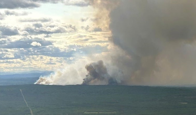 Aerial view of a large wildfire burning in a forested area, with thick smoke billowing high into the sky under a partly cloudy sky.