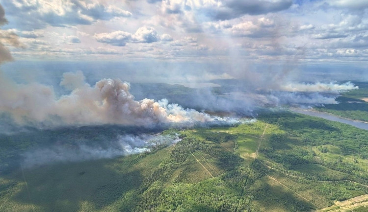 Aerial view of a wildfire burning through forested land. Thick smoke rises in multiple columns and drifts across the landscape under partly cloudy skies.
