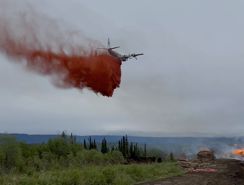 n airtanker drops red fire retardant over a wildfire burning in a log deck, with smoke rising from the flames below. The aircraft is flying low under cloudy skies, and forested hills are visible in the background.