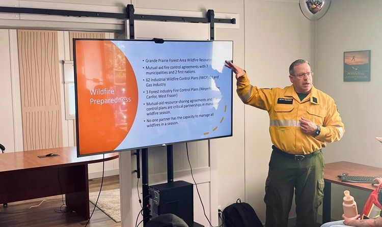A wildfire preparedness presentation is being delivered in a meeting room. A man in a yellow Alberta Wildfire uniform points to a large screen displaying key points about wildfire preparedness in the Grande Prairie Forest Area. 