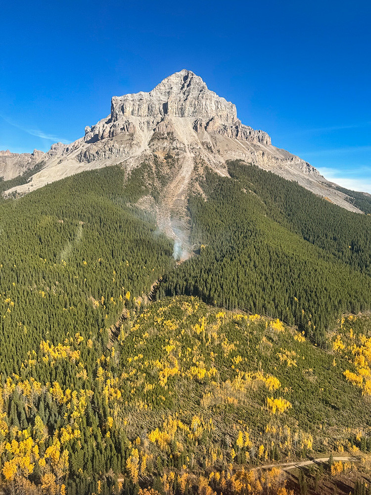 Mountain peak rising above a dense evergreen forest with patches of golden autumn foliage. A small plume of smoke is visible in the trees below the rocky summit under a clear blue sky.
