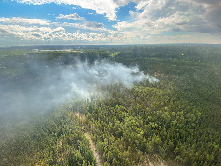 Aerial view of a forest with a smoke plume rising from an active wildfire among dense green trees. A narrow access road leads toward the hotspot, while clouds stretch across the sky above the remote landscape.