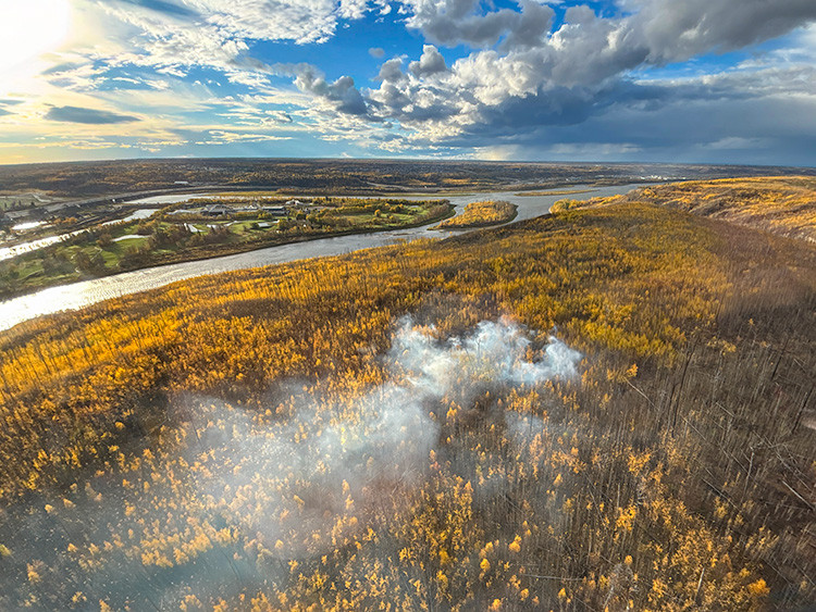 Aerial view of a small smoke plume rising from a forested river valley filled with golden autumn trees. A winding river cuts through the landscape beneath dramatic clouds and bright patches of sky.