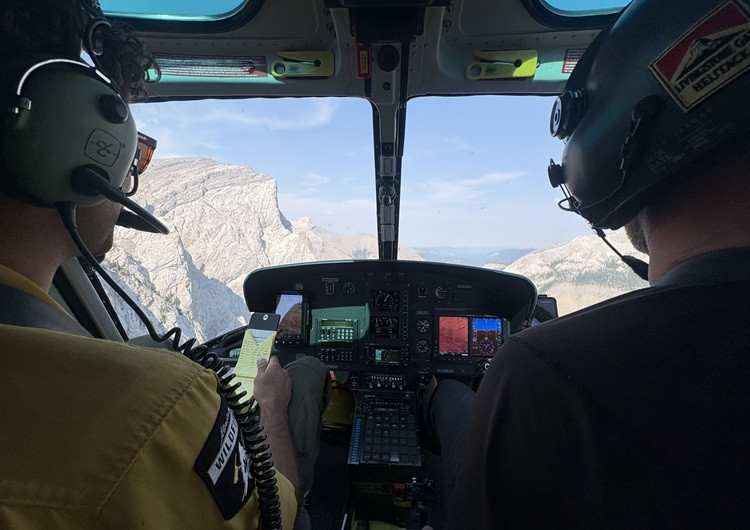 View from inside a helicopter cockpit during an aerial patrol, with two crew members wearing headsets and rugged mountain peaks visible through the windshield. Flight instruments and navigation displays are visible in the foreground.