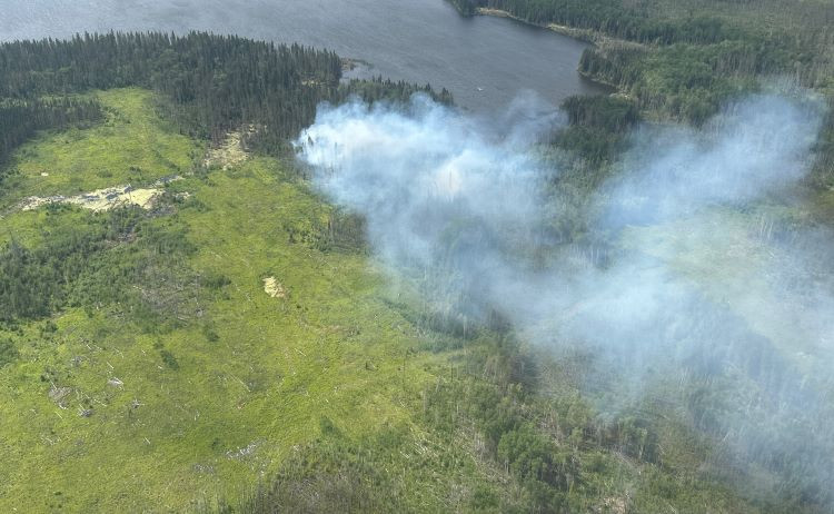 Aerial view of wildfire SWF059 burning near McConachie lake. Light gray smoke drifts across a mix of green vegetation, wetlands, and dense conifer trees with water visible in the background.