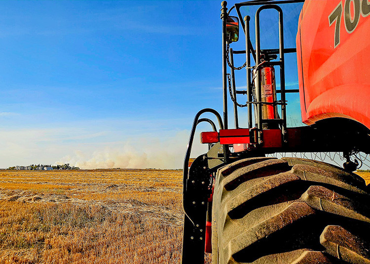 Close-up of a large farm machine tire in a harvested field under a clear blue sky. Smoke rises in the distance near a tree line, highlighting a potential grass or field fire on the horizon.