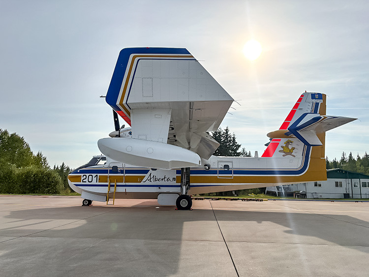 Alberta Wildfire CL215T air tanker parked on an airport tarmac. The white and yellow aircraft is backlit by the sun, with trees and airport buildings in the background.