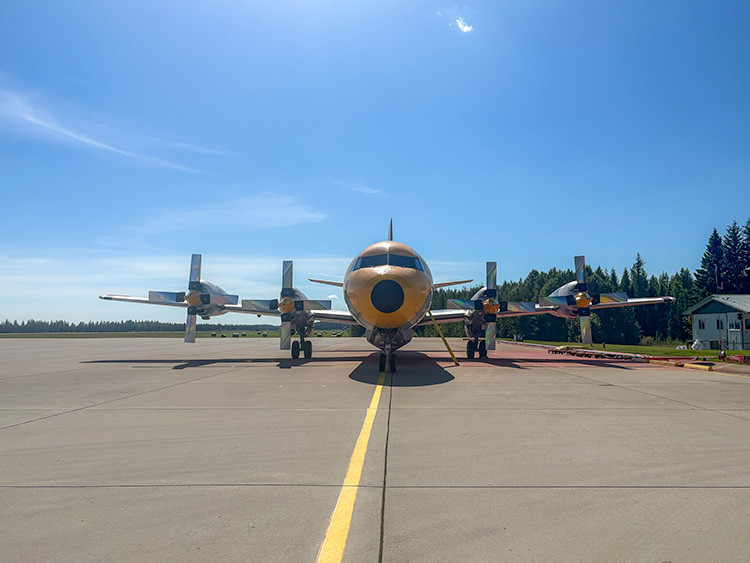 Front view of a twin-engine propeller aircraft parked on an airport apron under a clear blue sky. The aircraft is centered on a yellow taxiway line, with trees, small buildings, and fuel hoses visible in the background.