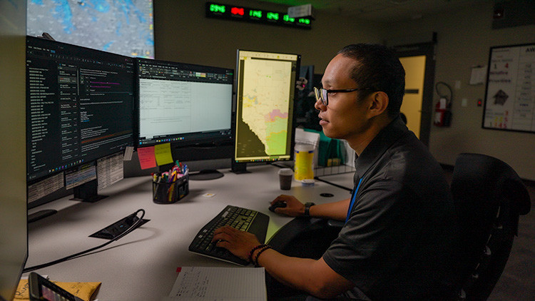 Person working at a multi-monitor computer workstation in an operations centre, viewing maps and software dashboards while using a keyboard and mouse. Digital status displays and office equipment are visible in the background.