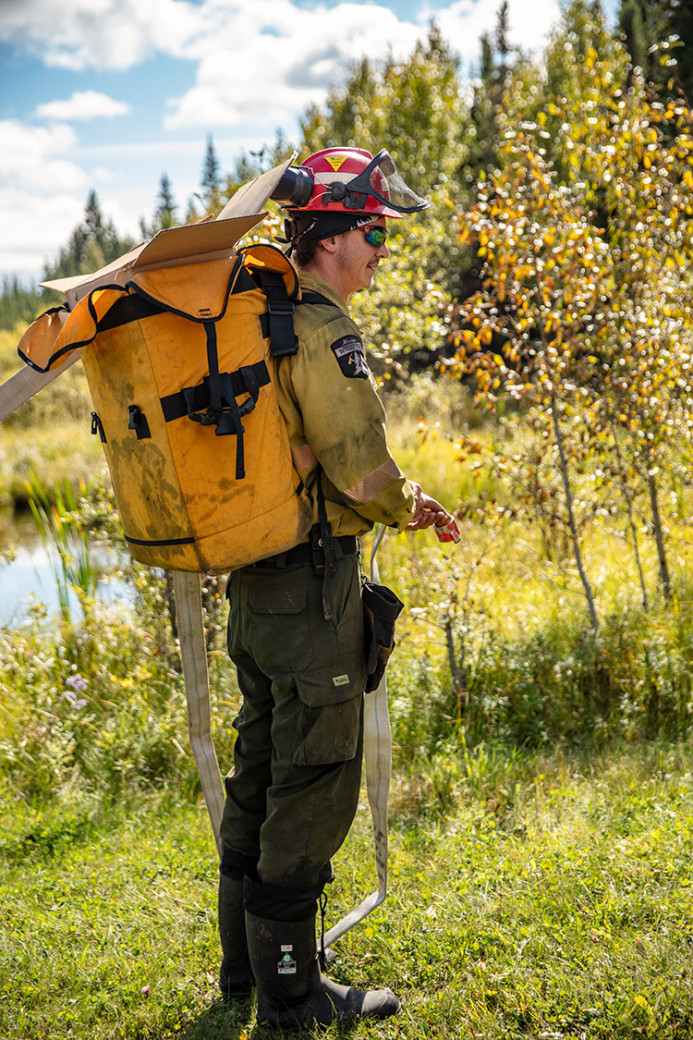 Wildfire worker in protective clothing and helmet carries a backpack hose pack while standing in a grassy area near a pond. Trees and shrubs surround the scene as the worker prepares firefighting equipment for field use.