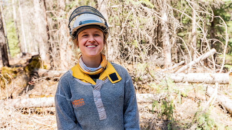 A wildfire worker wearing a hard hat and protective clothing stands in a forested area with fallen logs and trees in the background. The worker is facing the camera in a sunlit woodland setting.
