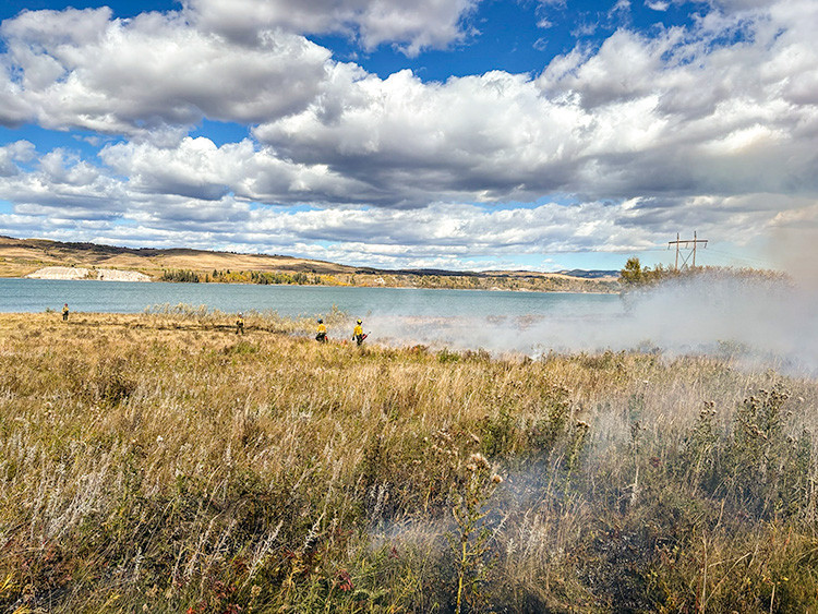 Firefighters conduct a hazard reduction burn beside a lake. Light smoke drifts across dry vegetation as crews monitor the burn under a dramatic sky filled with large clouds.