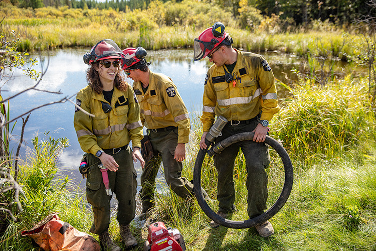 Three wildfire personnel in protective gear and helmets work beside a pond during pump operations. One holds a hose while others stand near equipment at the water’s edge, surrounded by tall grass and forest.