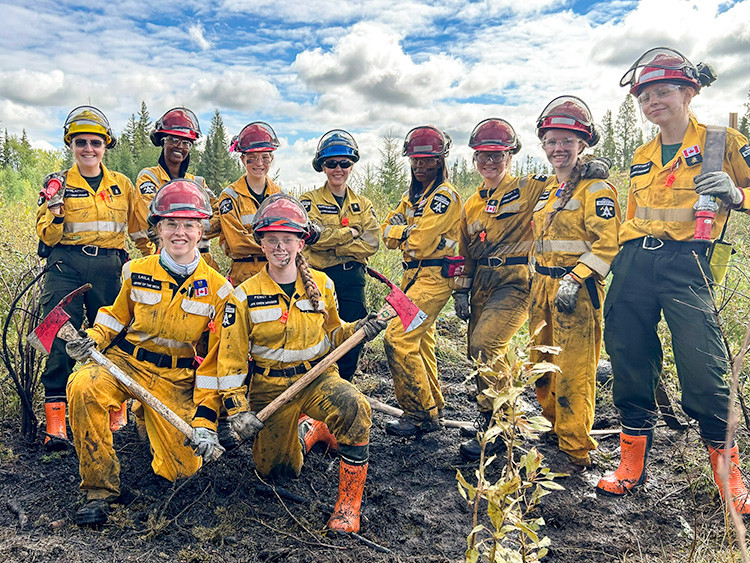 Junior Forest Ranger crew members in yellow protective gear and hard hats pose after mock fire exercise, holding hand tools. Charred ground, young trees, and a cloudy sky are visible in the background.