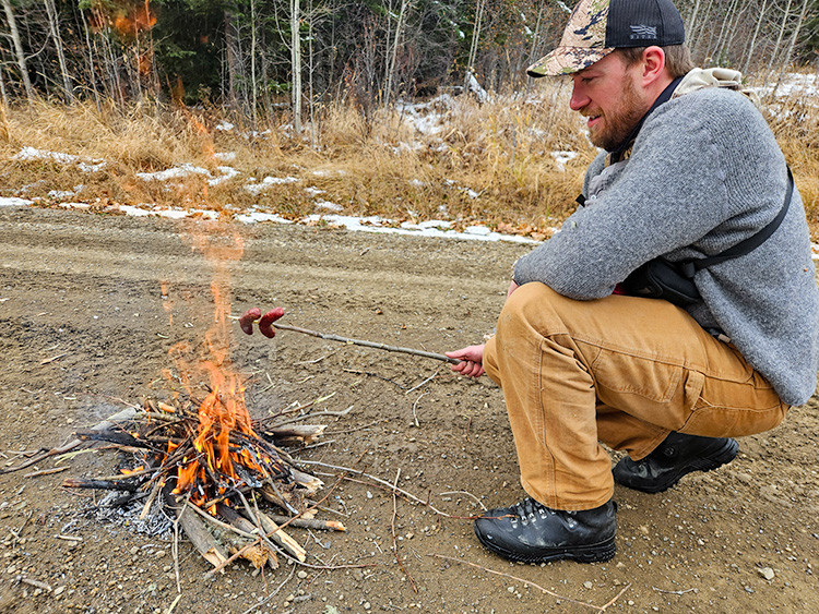 A person crouches beside a small campfire on a gravel roadside, holding a roasting stick over the flames. Dry grass, patches of snow, and a forested backdrop surround the scene.