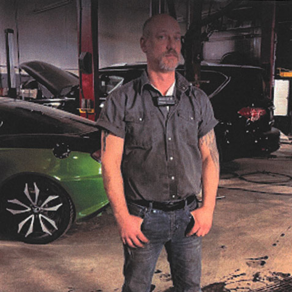  Eric Charles Andrew Janzen: Bald white male with a mustache and goatee, standing in a mechanic shop wearing a blue buttoned shirt and jeans, green and black vehicles in the background